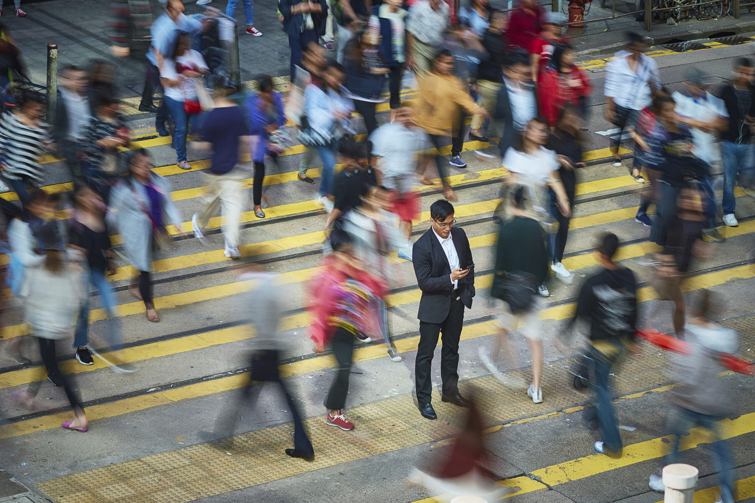 Motion-blurred view of people in a busy crosswalk. A single businessman stands stationary in the middle.