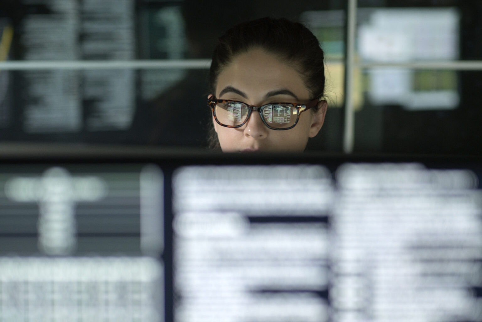 Woman with glasses working at a bank of computer monitors.