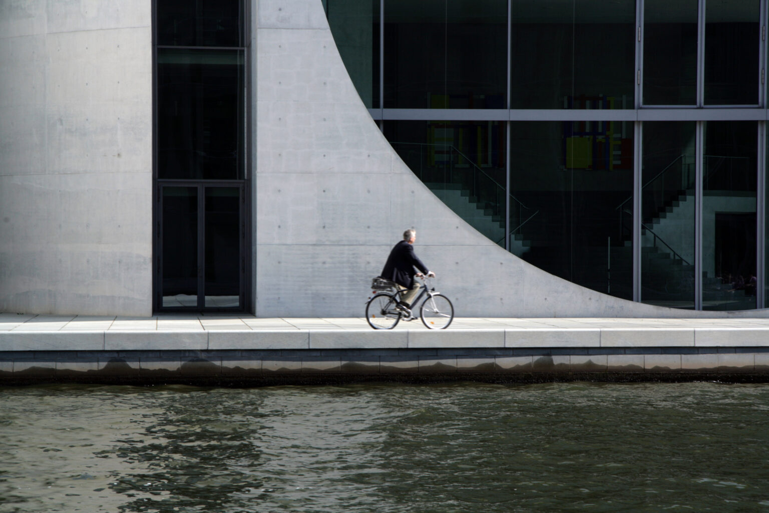 Artistic shot of a businessman on a bicycle with curved architectural wall in the background.