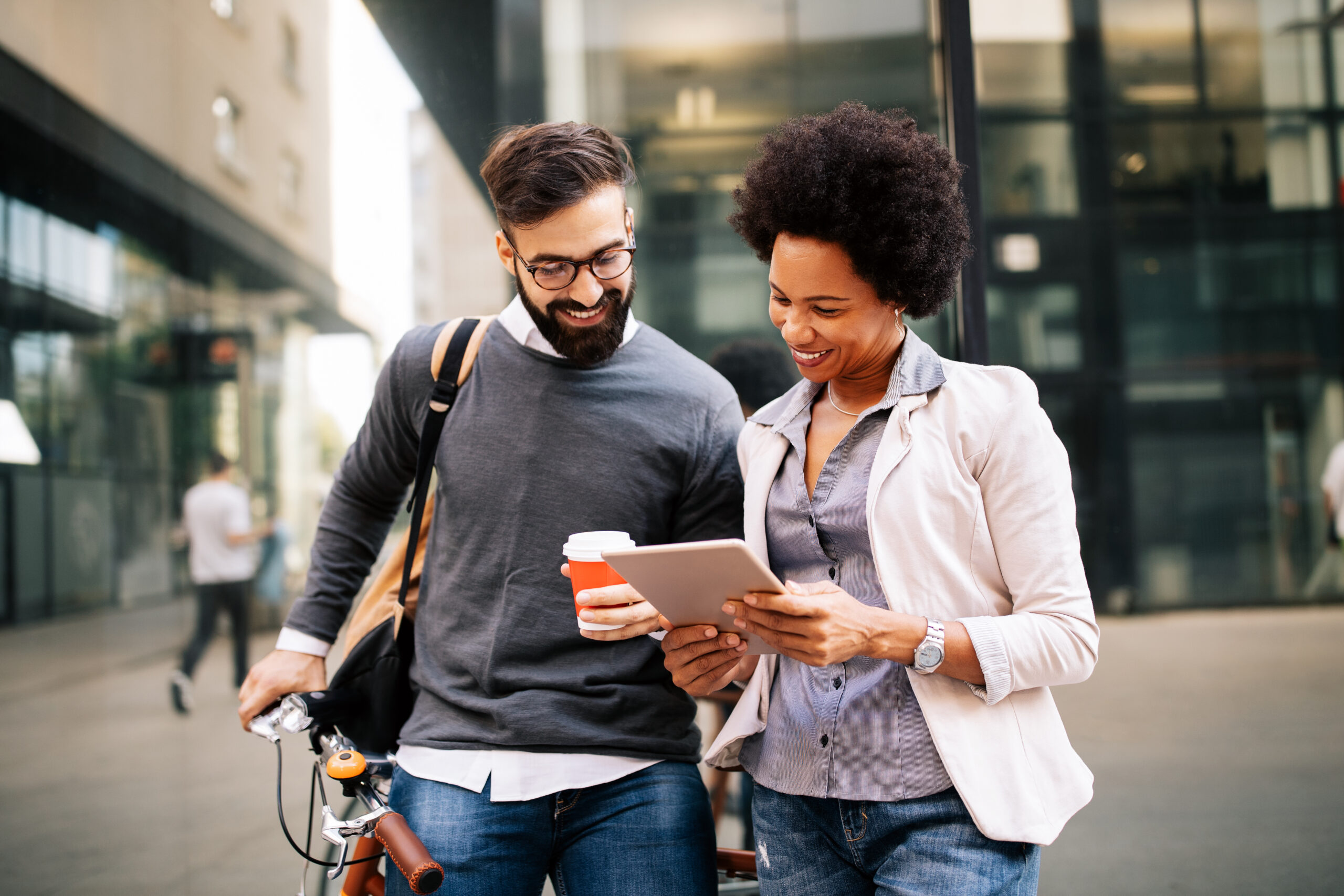 A smiling man with a bike and a cup of coffee talking with a smiling woman with a tablet computer.