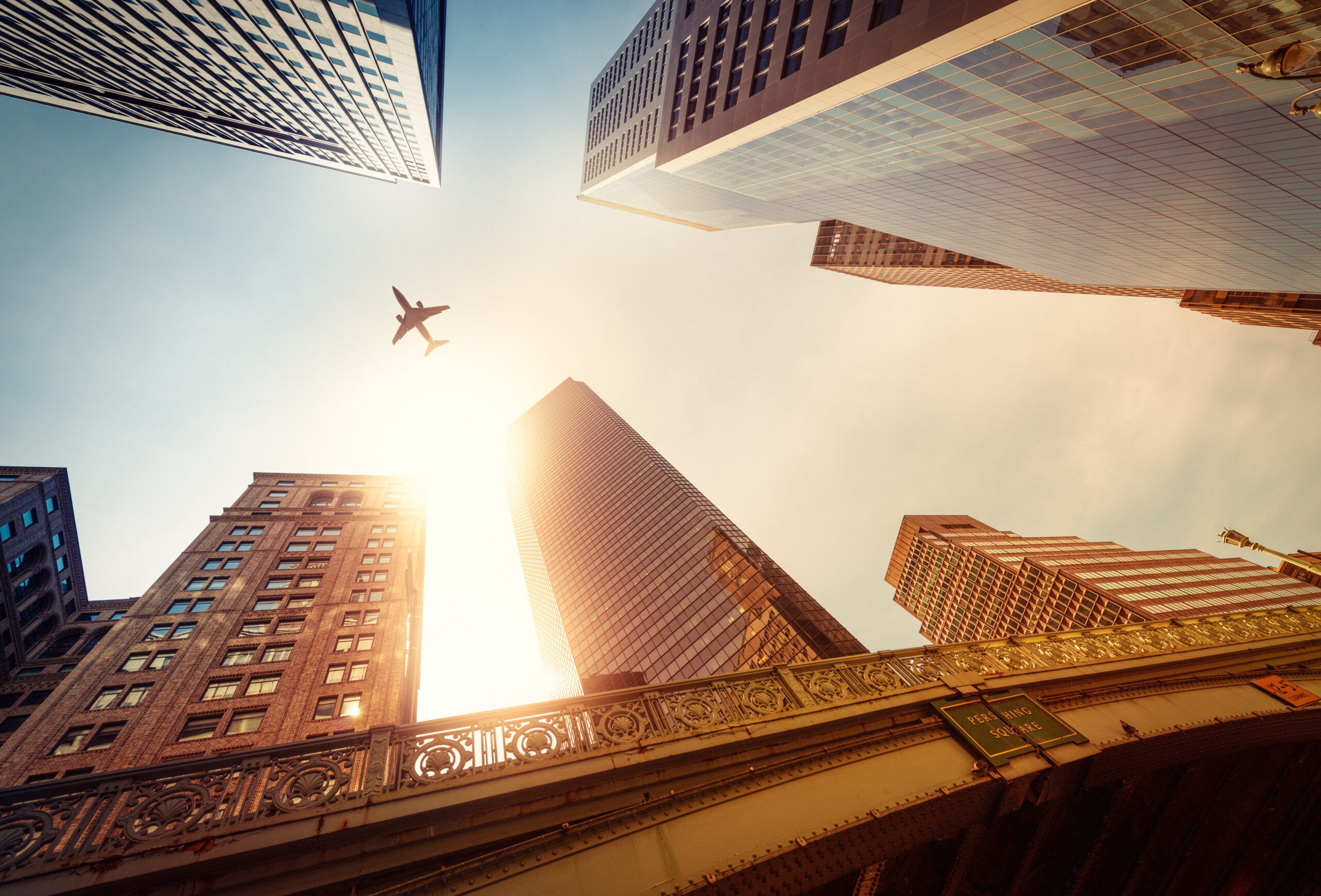 A skyward view of buildings at sunset with a jet plane high above.