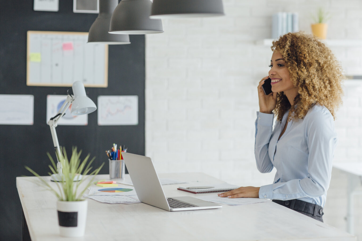 Smiling woman at a desk working on a computer and talking on a mobile phone.