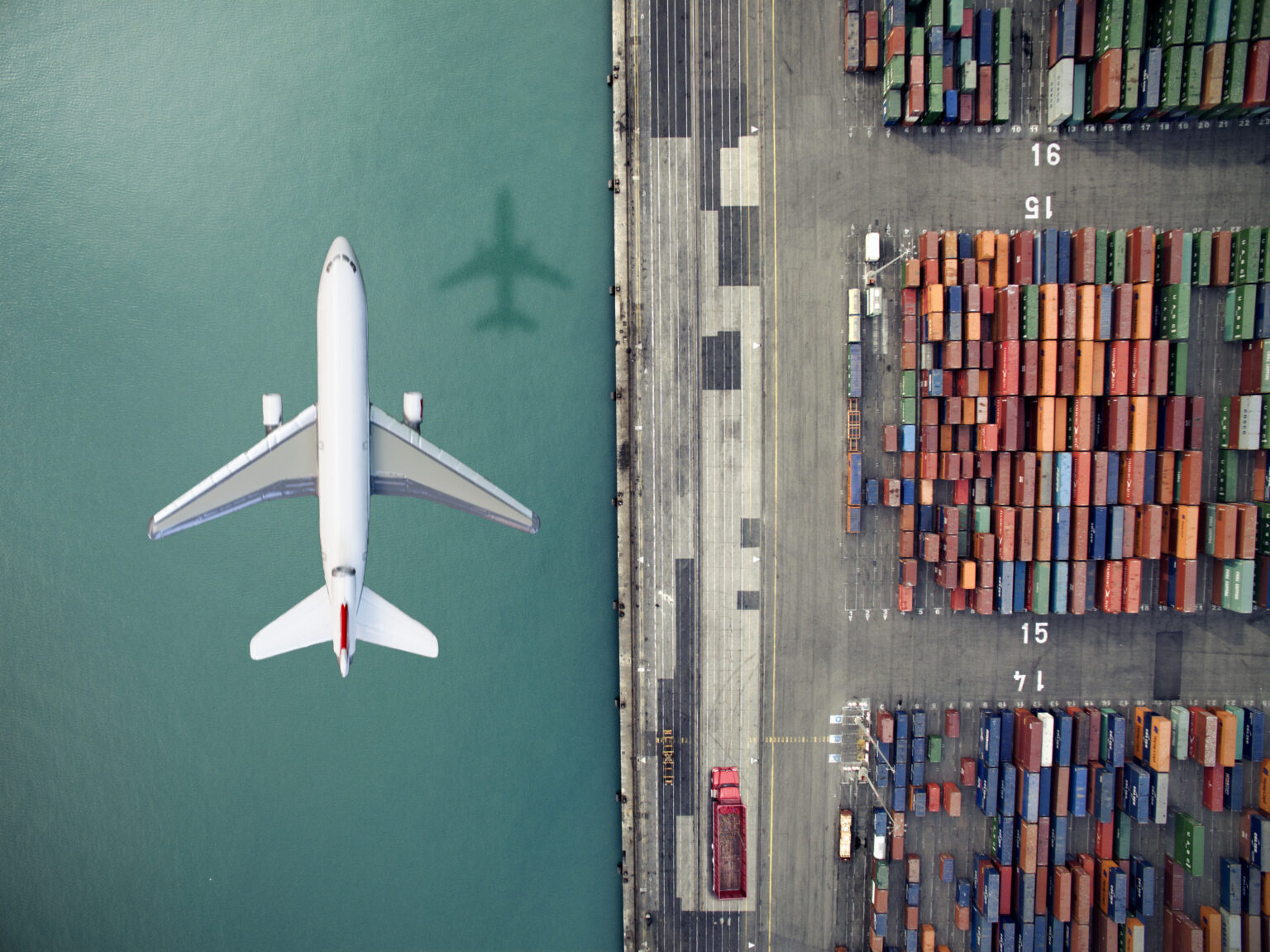 An overhead view of a jet plane flying over a waterway with colorful shipping containers stacked nearby.