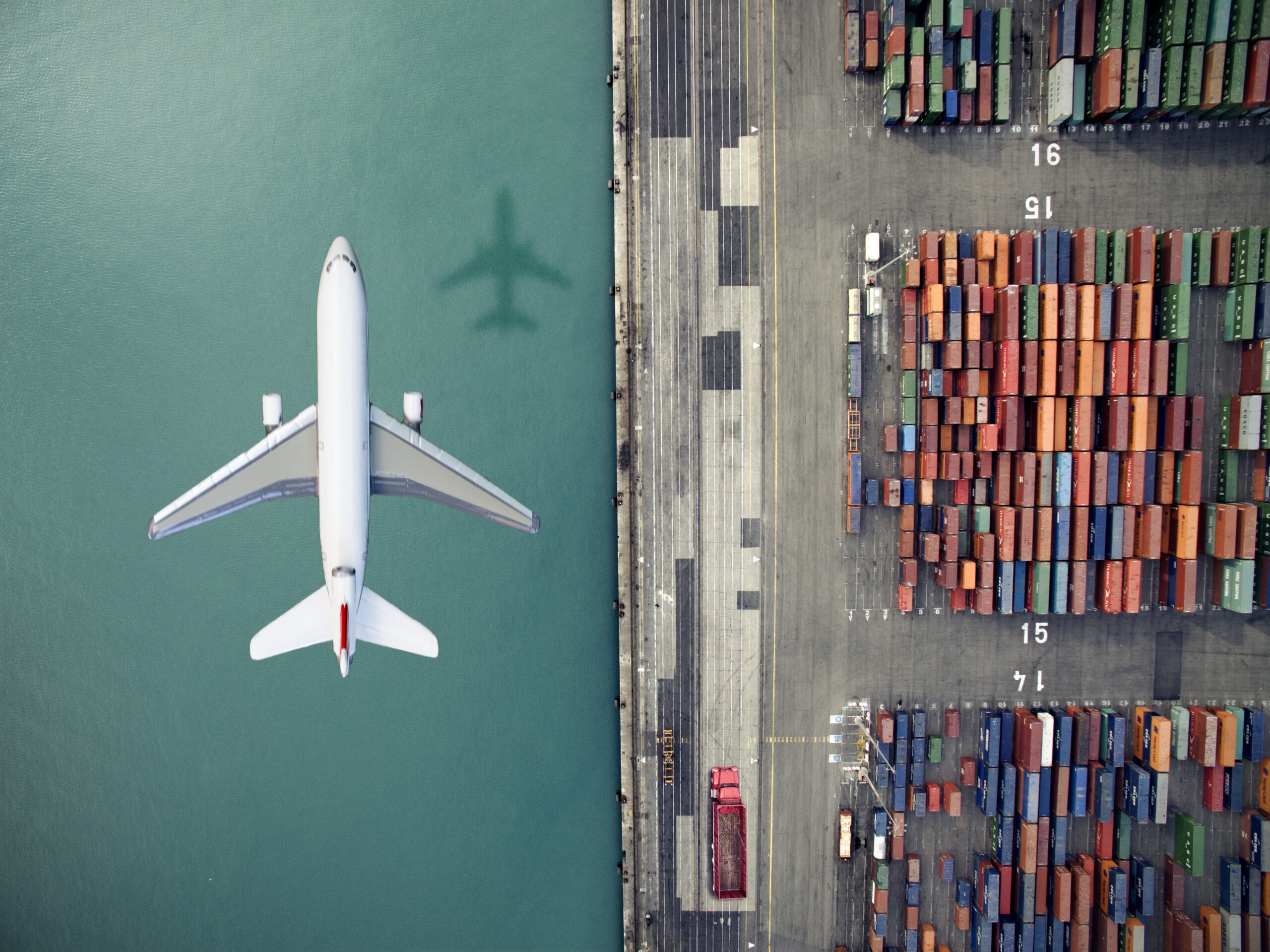 An overhead view of a jet plane flying over a waterway with colorful shipping containers stacked nearby.