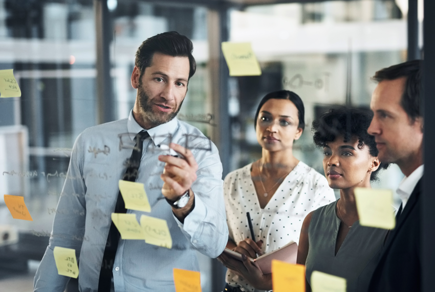 A group of four professionals gathered around a glass wall covered in sticky notes, discussing ideas. The man in the foreground is pointing at the notes, while the others listen attentively, taking notes.