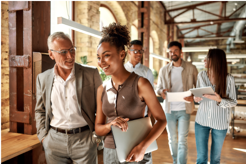 A diverse group of professionals walking in a modern office, discussing work, holding laptops and documents, in a collaborative, industrial-style setting.
