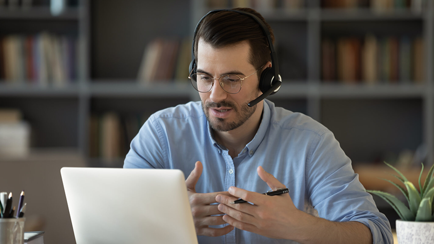 A man with a headset on gesturing while working on a laptop computer.